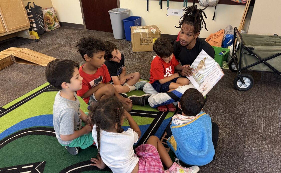 Tr Seth is reading a story to his class on the rug. They're all leaning toward him and he's asking them a question.